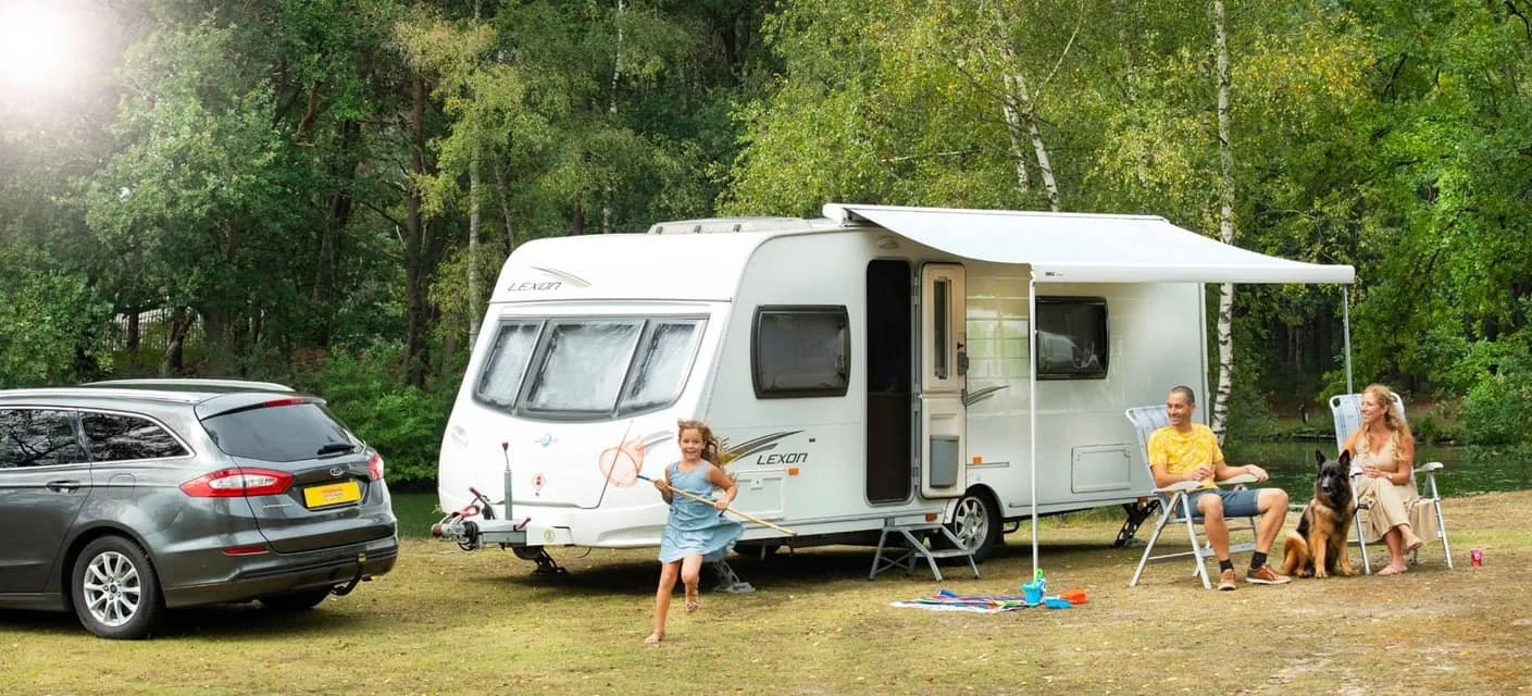 Gezinnen genieten van een campingveld met een witte caravan en een geparkeerde auto. Een meisje danst met een hoepel naast de caravan, terwijl een man en een vrouw met een Duitse herdershond onder een uitklapbare luifel zitten en praten.