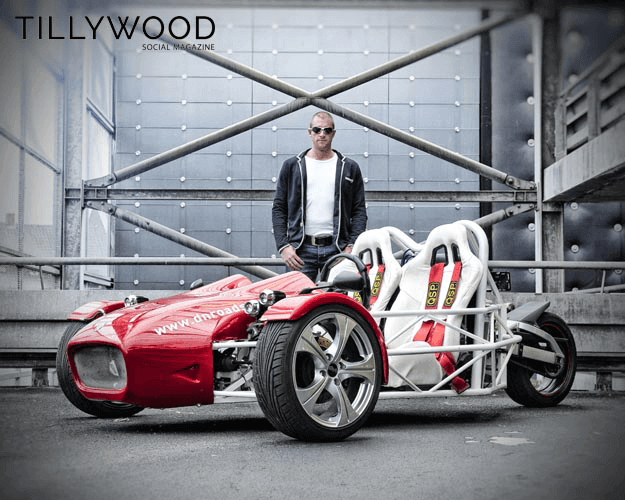 Man staat naast een lichtrode, open sportauto met twee racezetels, op een stedelijke brug met metalen reling en muur.