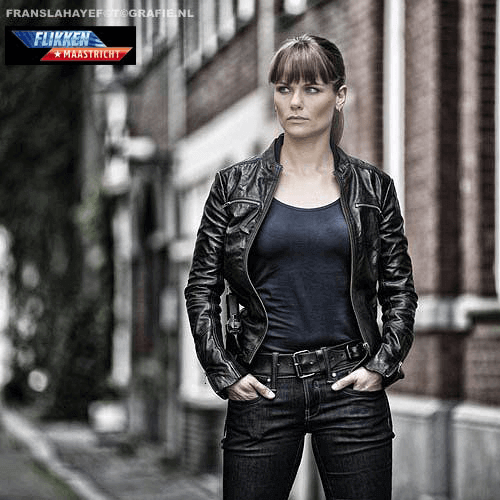 Een vrouw staat in een stadsgezicht met bakstenen gebouwen, gekleed in een zwarte leren jas en een blauw shirt.