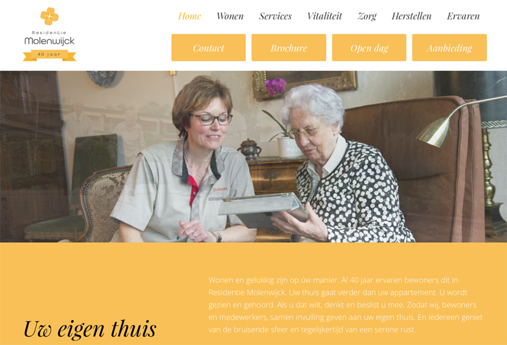 Een verzorgingshuis met een jonge vrouw die een oudere vrouw helpt met een tablet. Ze glimlachen samen in een gezellige, huiselijke omgeving.