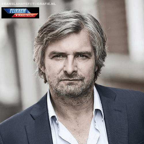 Man met grijze haar en baard, dragende een navyblauwe jas en wit overhemd, buiten met een bouwmuur op de achtergrond.