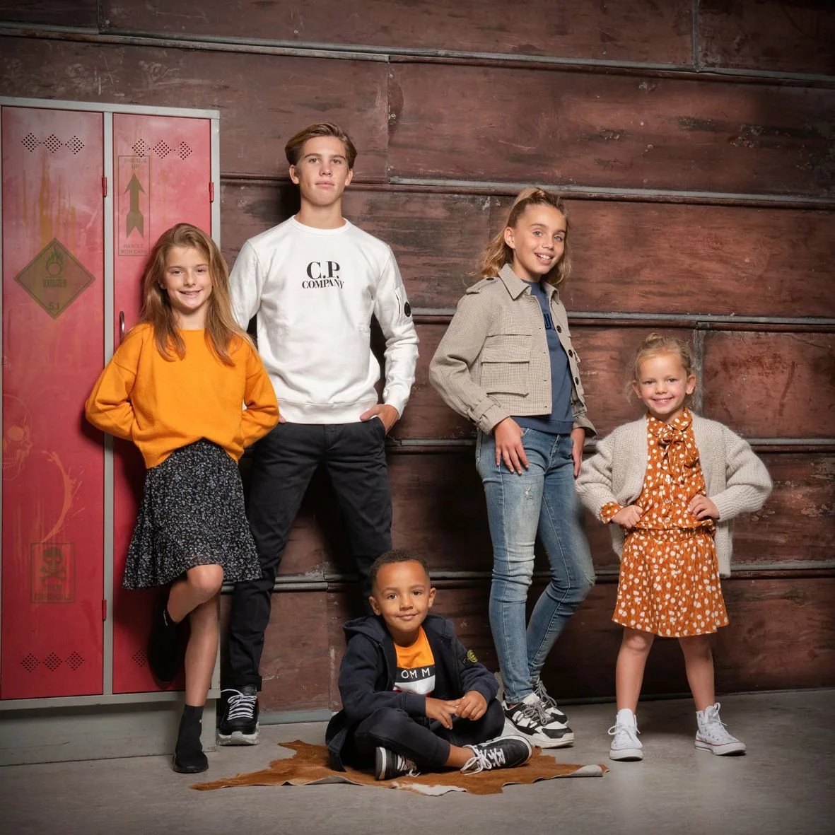 Vijf kinderen poseren in een kamer met een houten muur en rode kluisjes.