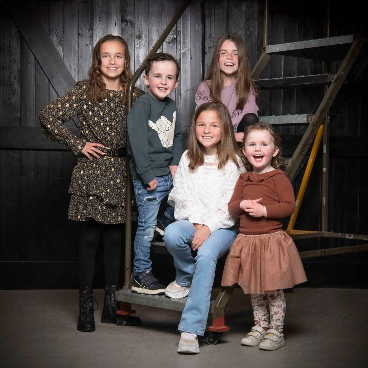 Vijf lachende kinderen poseren in een donkere houten kamer, met trappen en houten muren op de achtergrond.