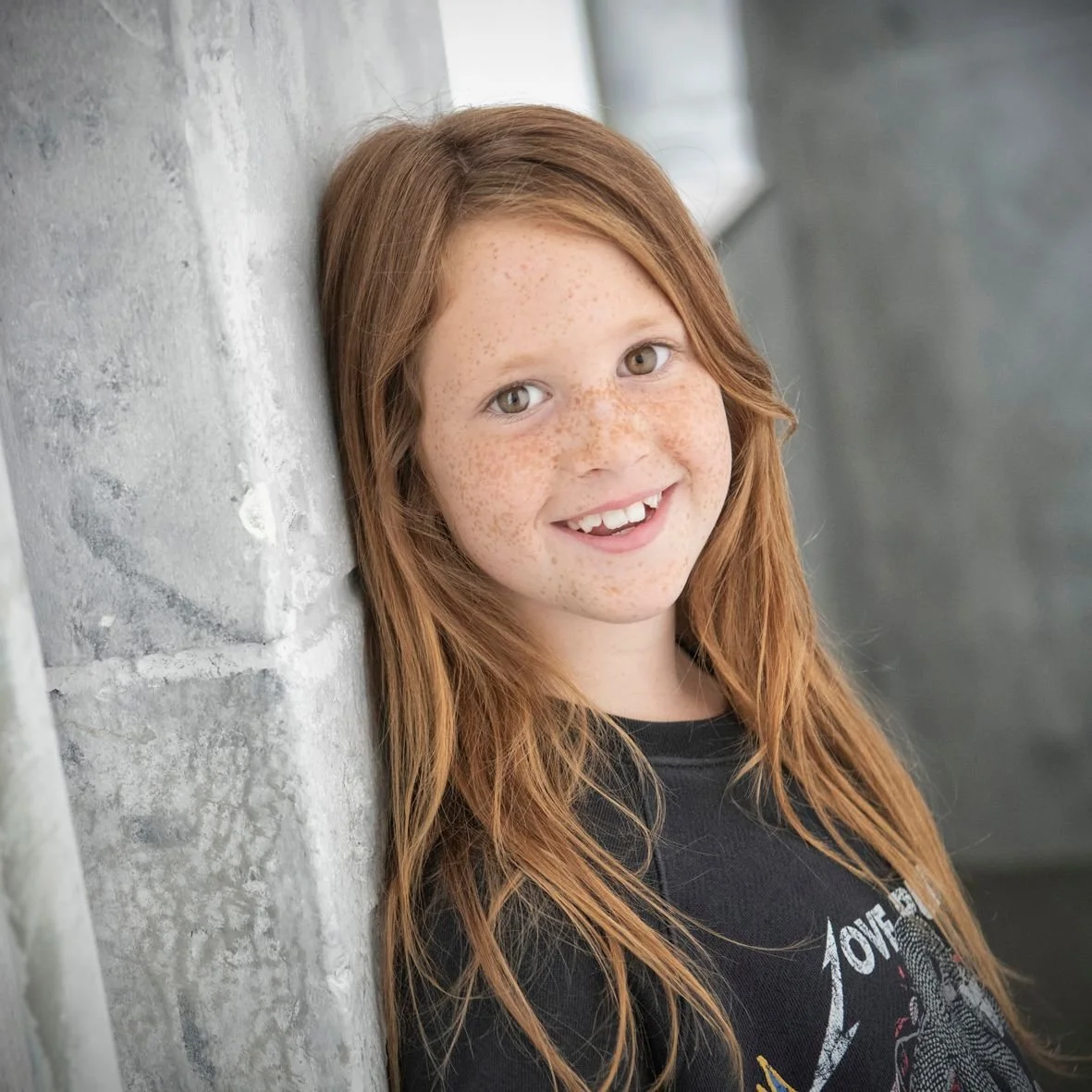 Een lachende roodharige jongen met sproeten, zittend tegen een bakstenen muur.