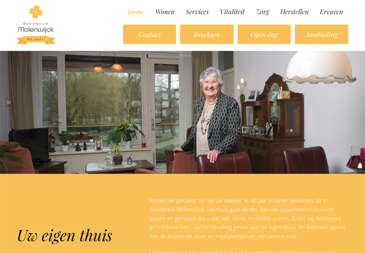 Een oudere vrouw staat glimlachend in een gezellig ingericht woonkamergedeelte, met grote ramen en tuinzicht.