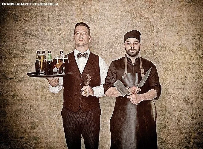 Twee mannen, een serveerster met dienblad met dranken en een chef-kok met messen, staan tegen een effen muur.