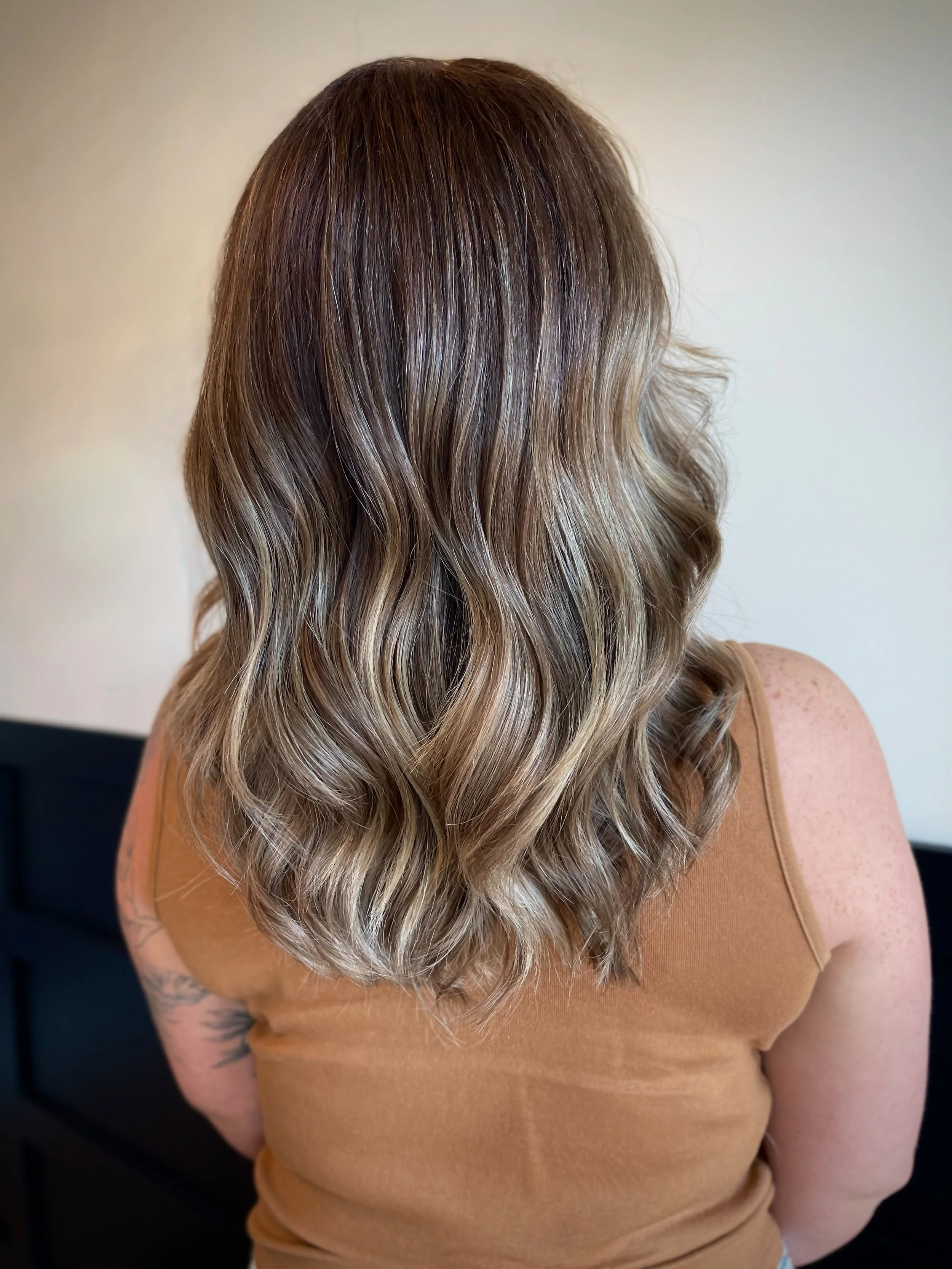 Back view of a woman with shoulder-length wavy blonde hair wearing a tan sleeveless top.