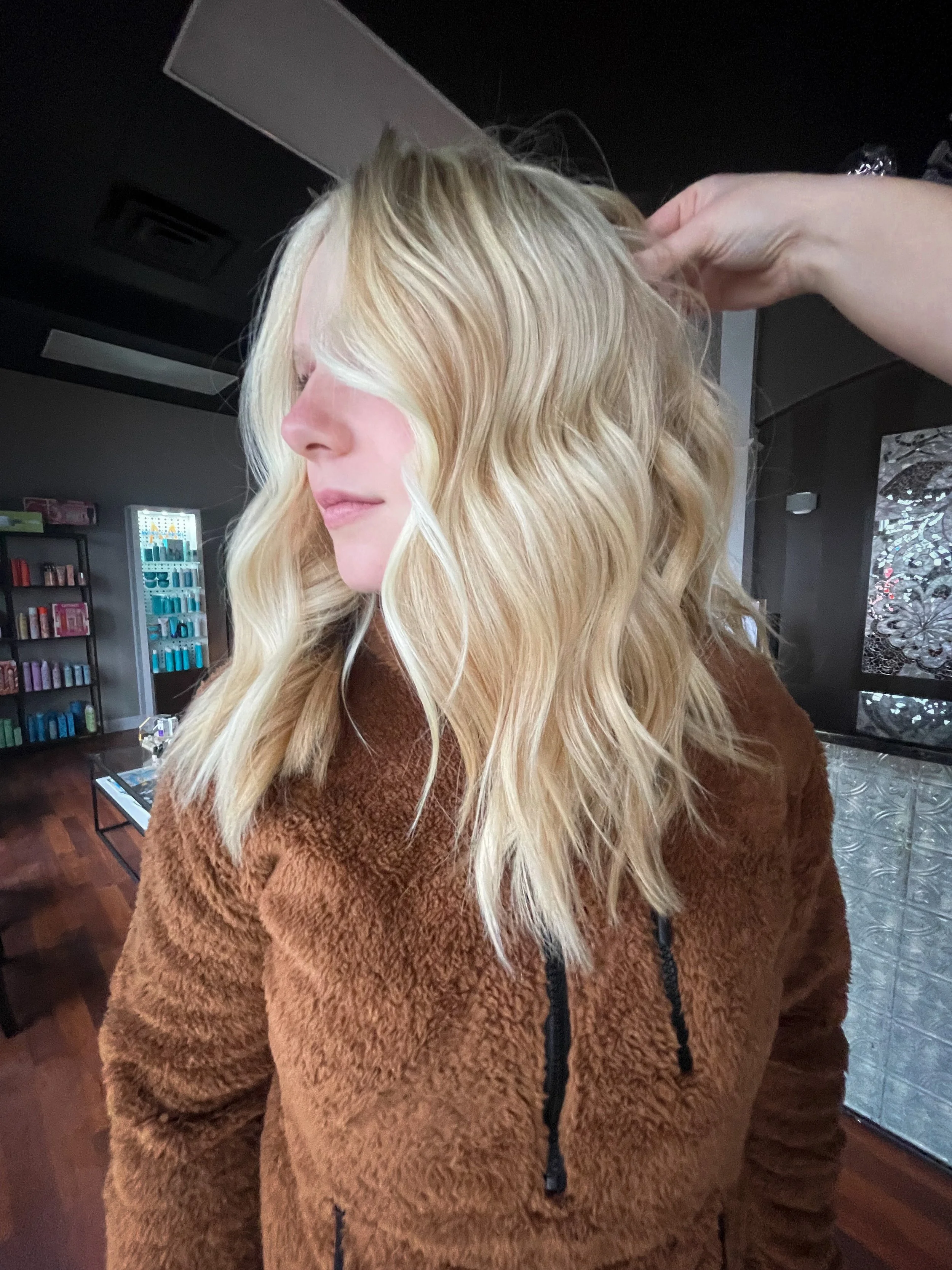 A woman with blonde, wavy hair in a salon, wearing a brown, fuzzy jacket.