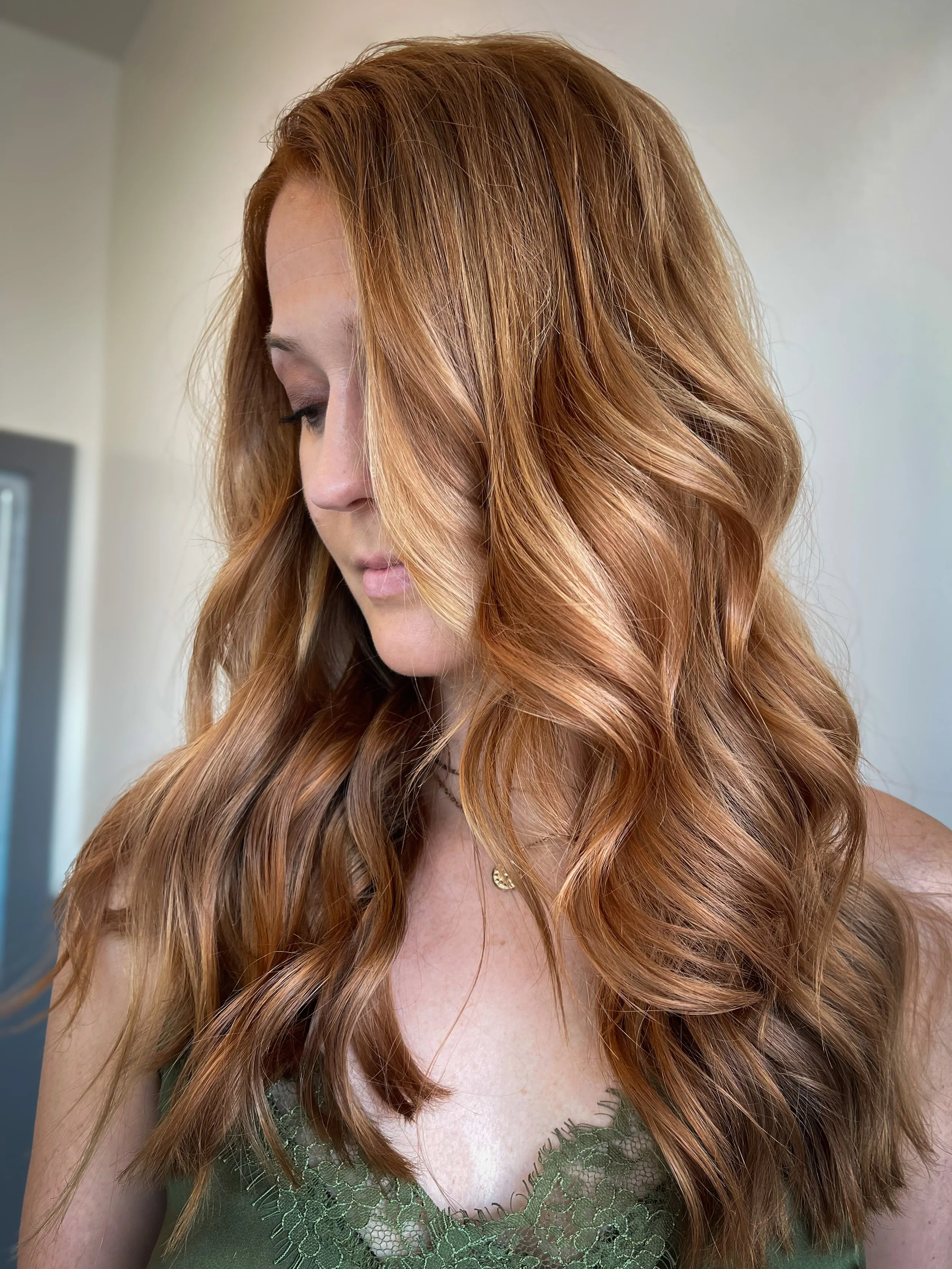 A woman with long, wavy red hair looking down, wearing a green lace top and a small gold necklace.