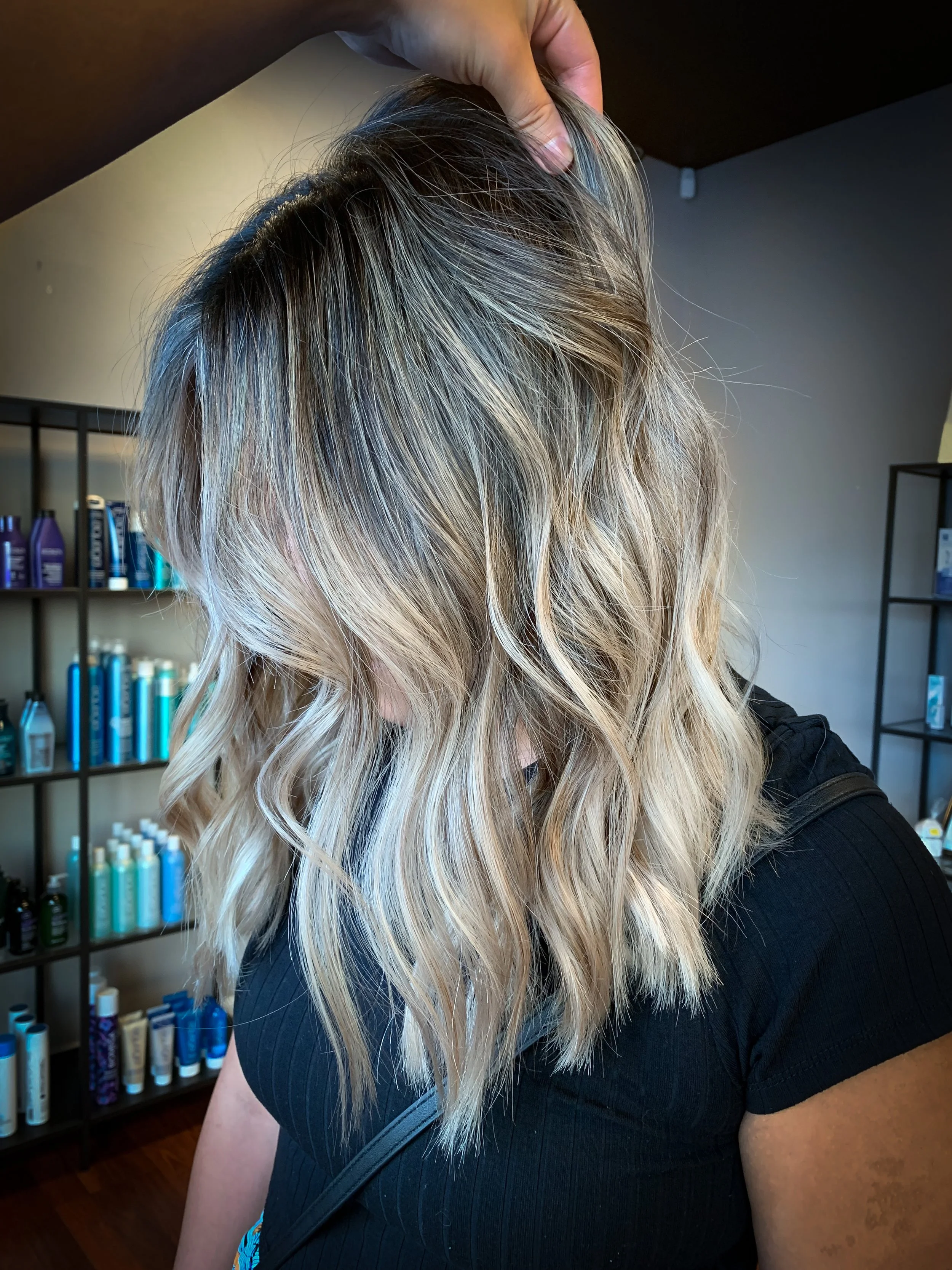A woman with shoulder-length, wavy blonde hair with darker roots, inside a salon with shelves of hair products in the background.