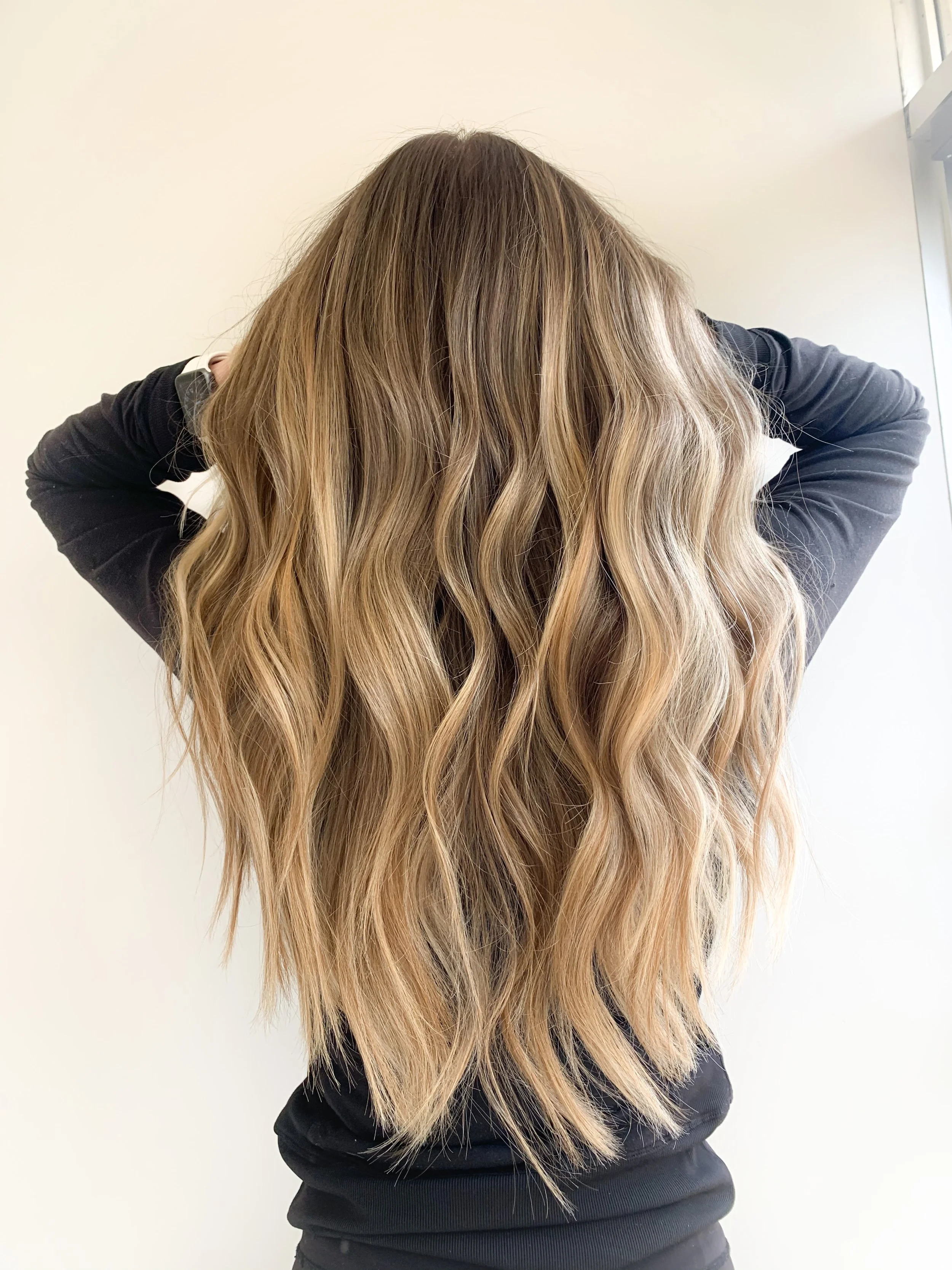 Woman with long, wavy blonde hair back view, wearing black long-sleeve top, standing against a plain background.