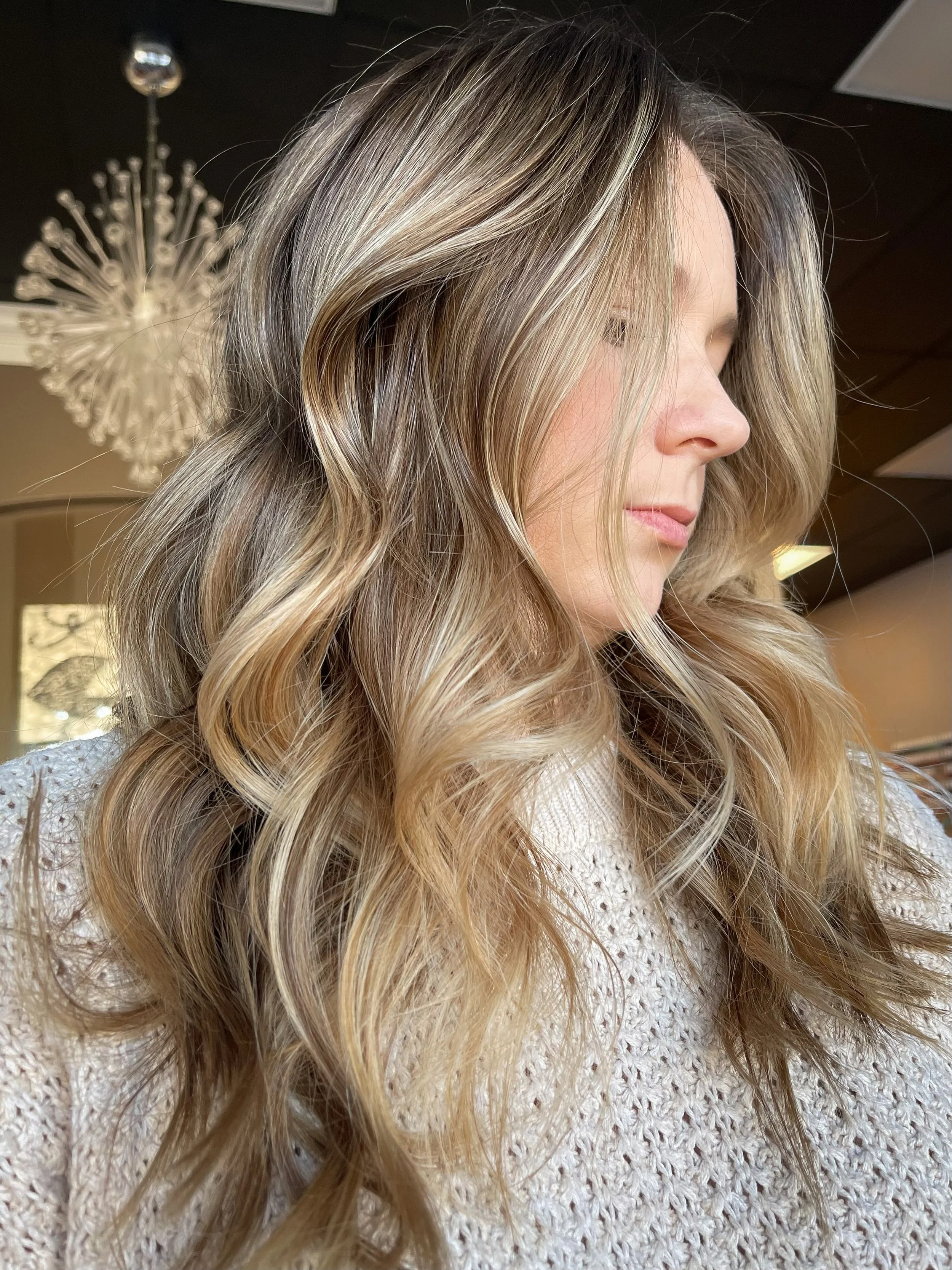 A woman with long, voluminous, curly blonde hair standing indoors, with a chandelier and warm lighting in the background.