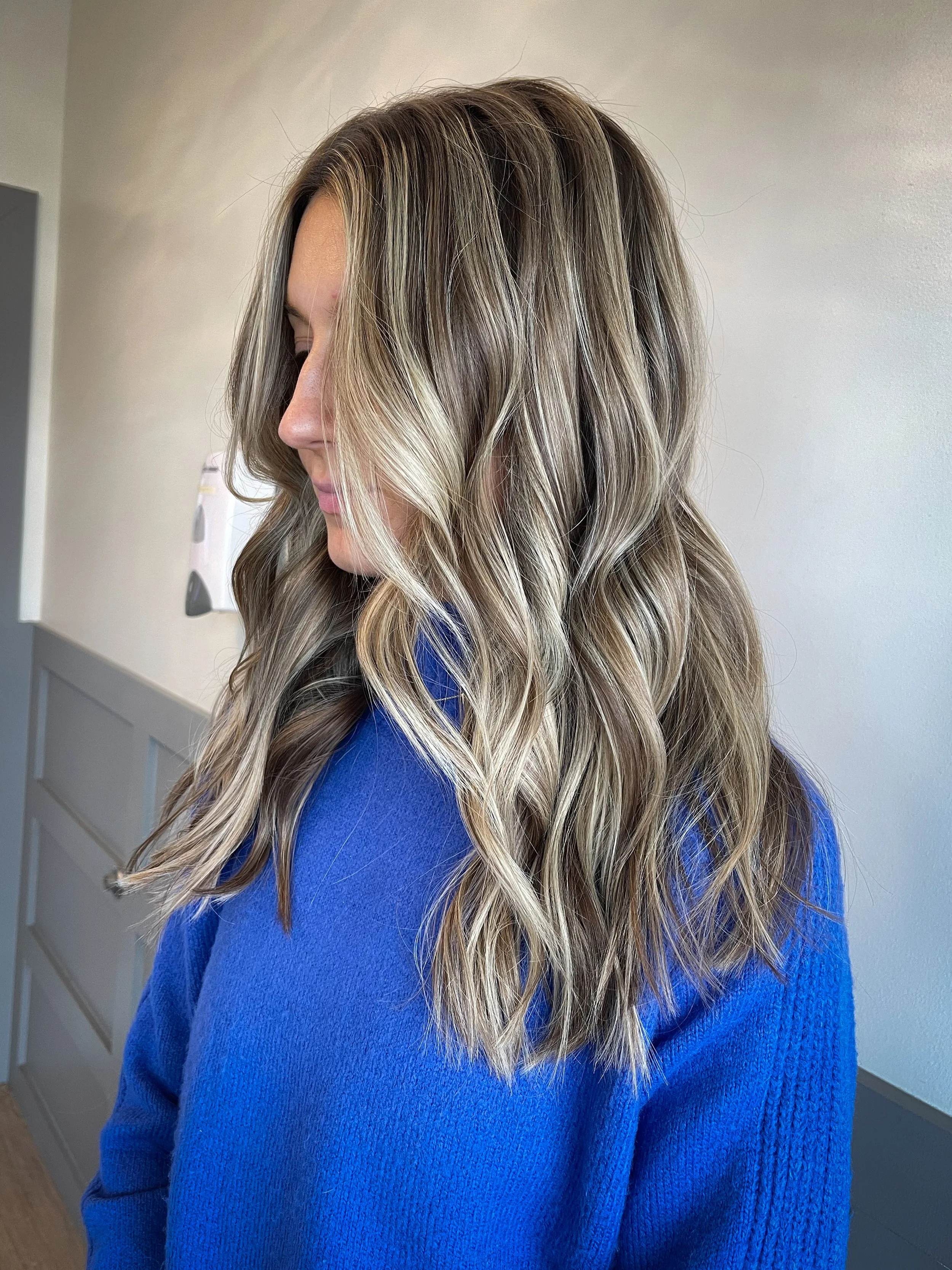 A woman with wavy blonde hair wearing a royal blue top, viewed from the side in an indoor setting.