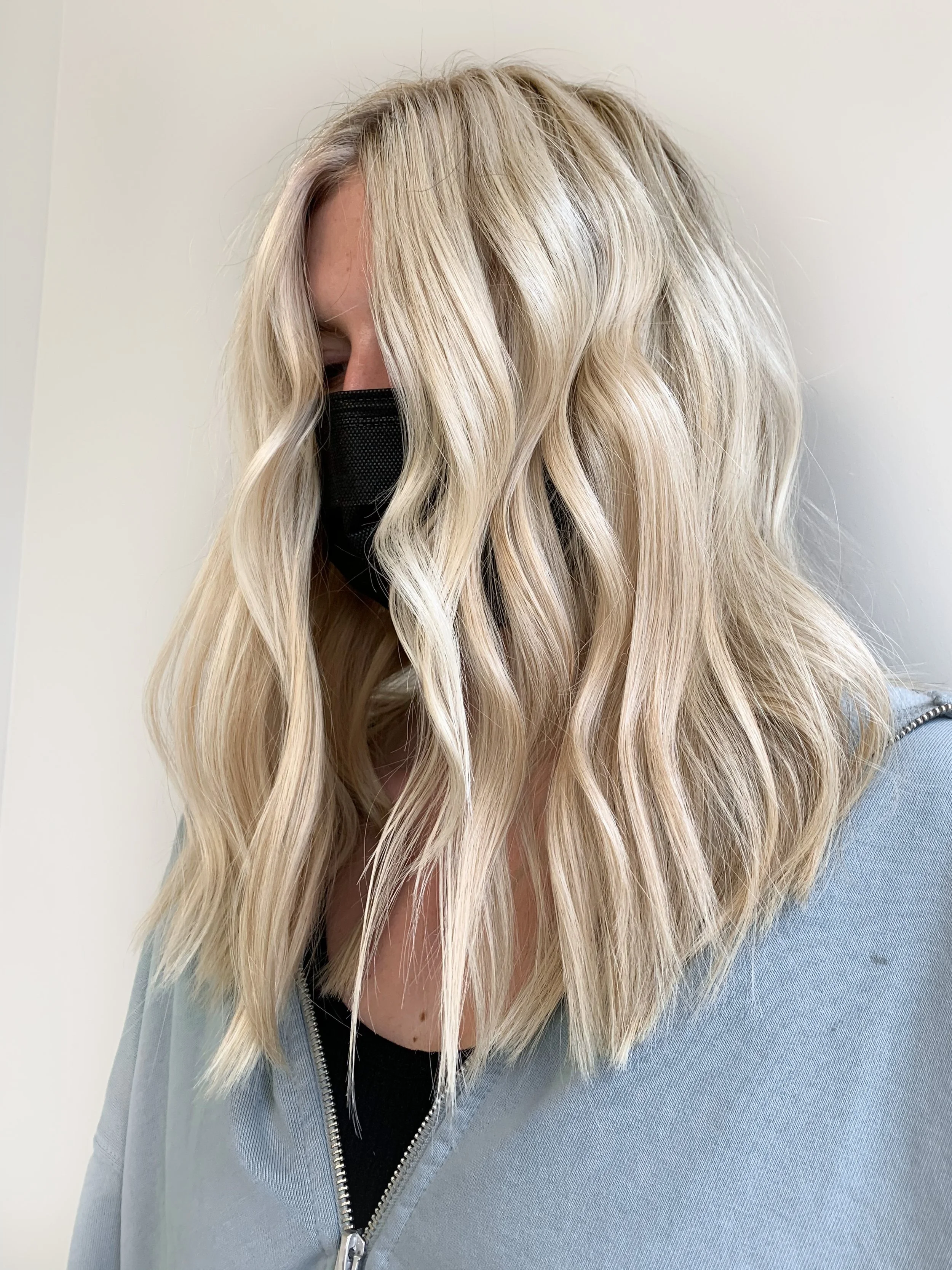A woman with wavy blonde hair wearing a black face mask and a light gray zip-up hoodie.