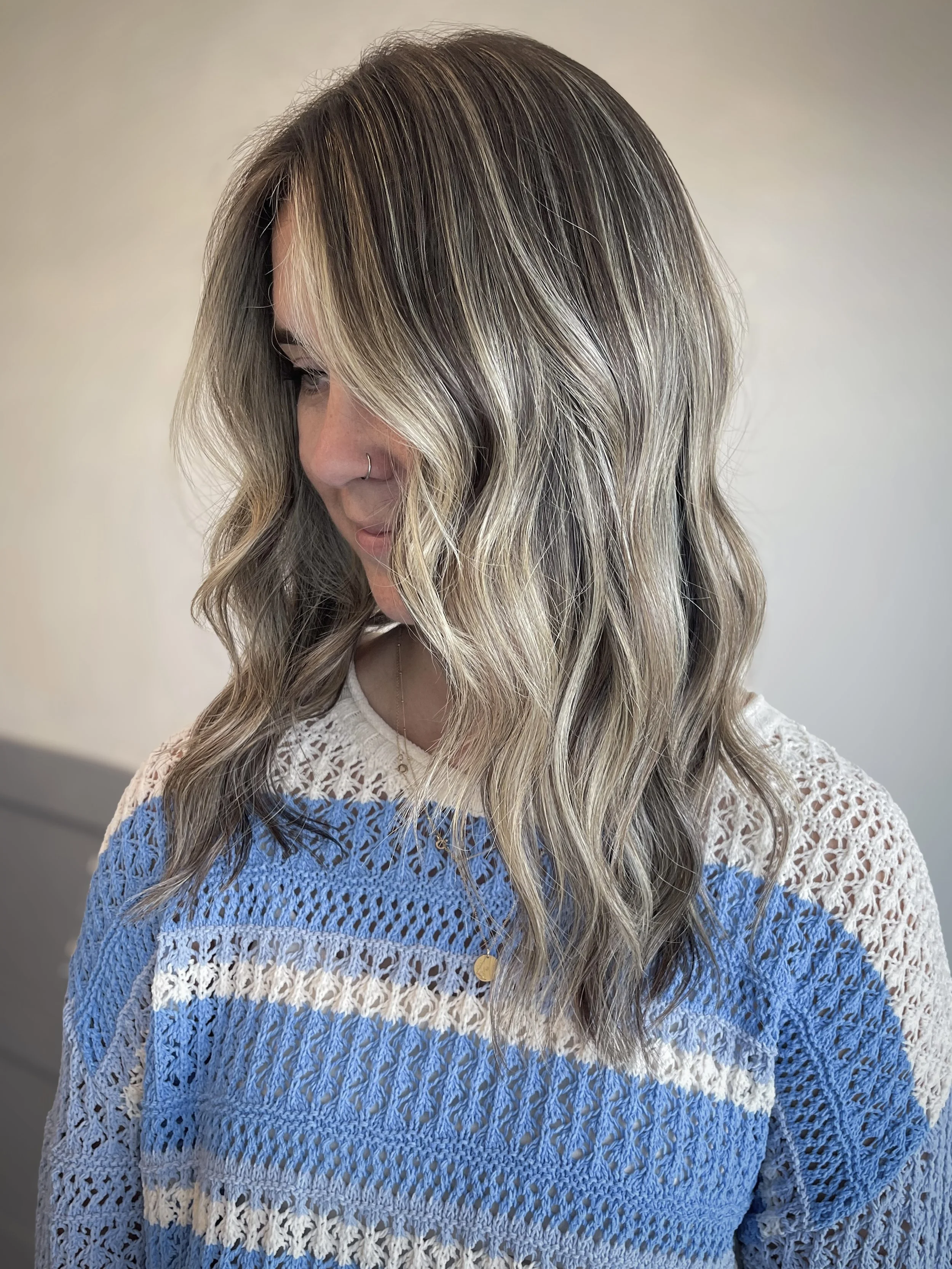 A woman with wavy, multi-toned blonde hair looking down, wearing a cream and blue crochet top with a gold necklace and nose ring.
