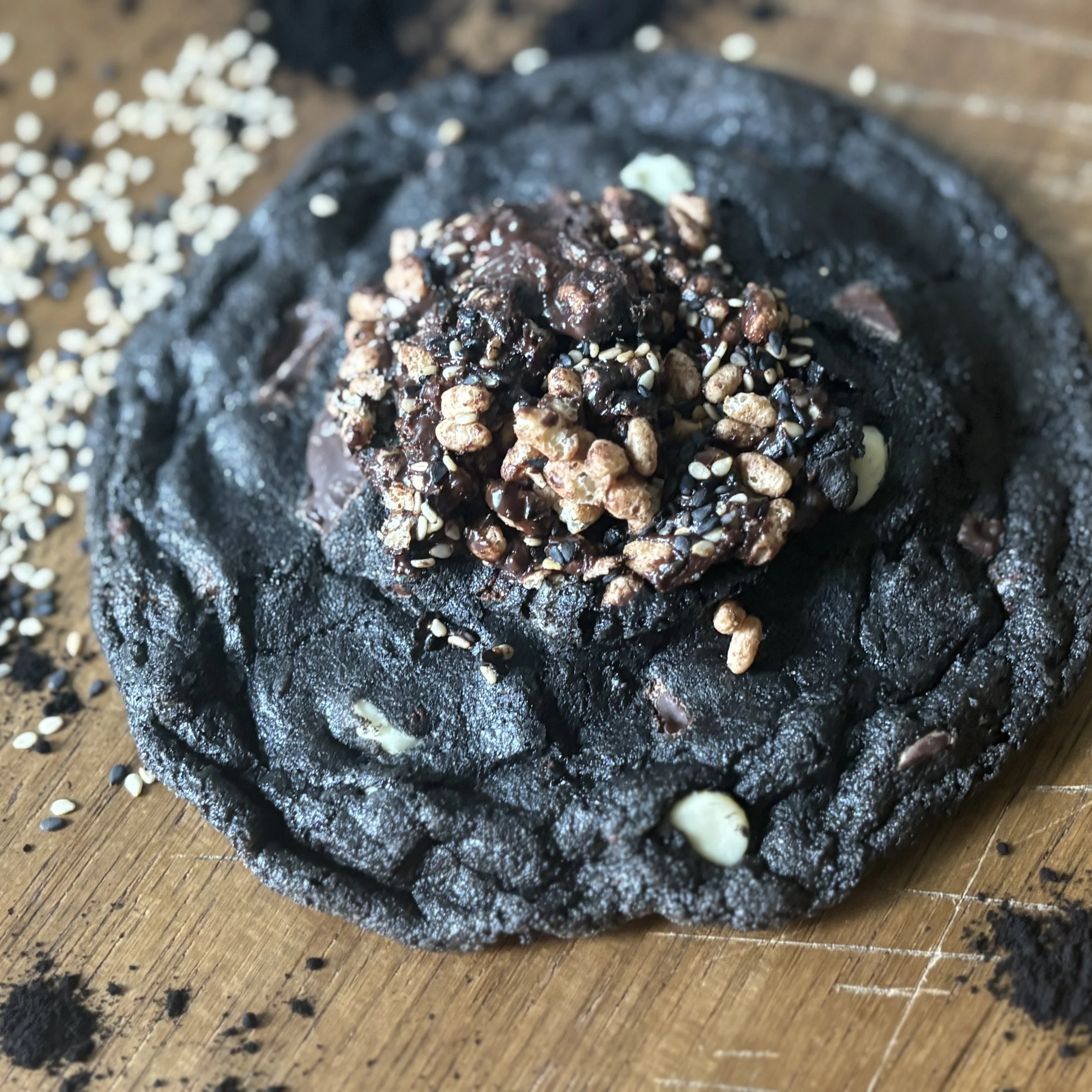 Close-up of a Chocolate black sesame seed cookie with sesame seeds on top, on a wooden surface with scattered sesame seeds.