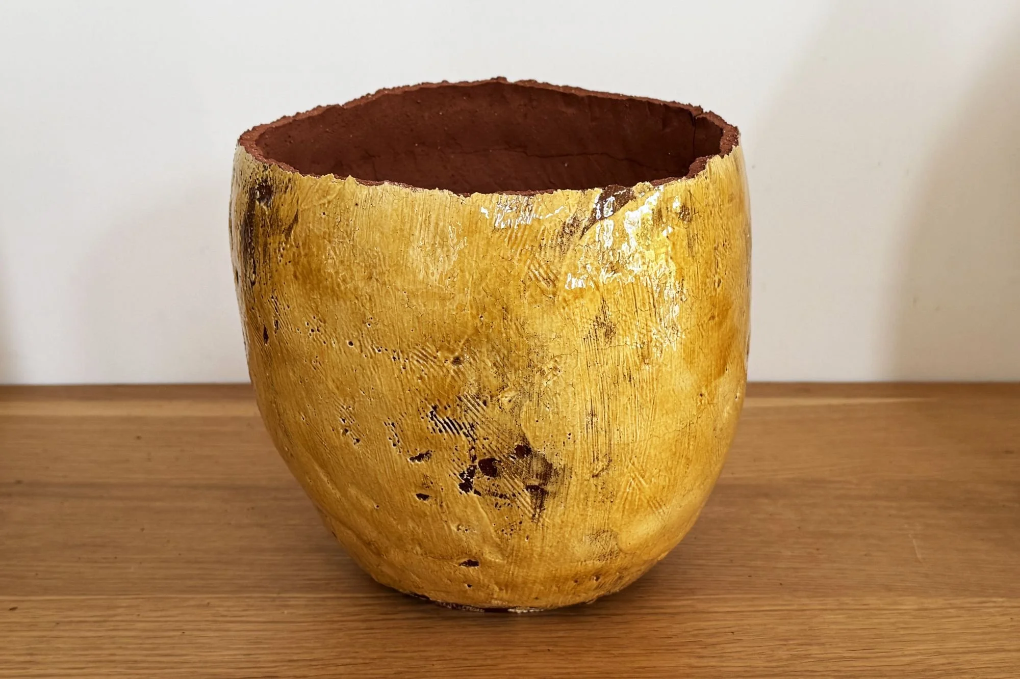 A large, hollow, yellow bowl with a rough, textured surface and a reddish-brown interior, placed on a wooden surface against a plain white background.