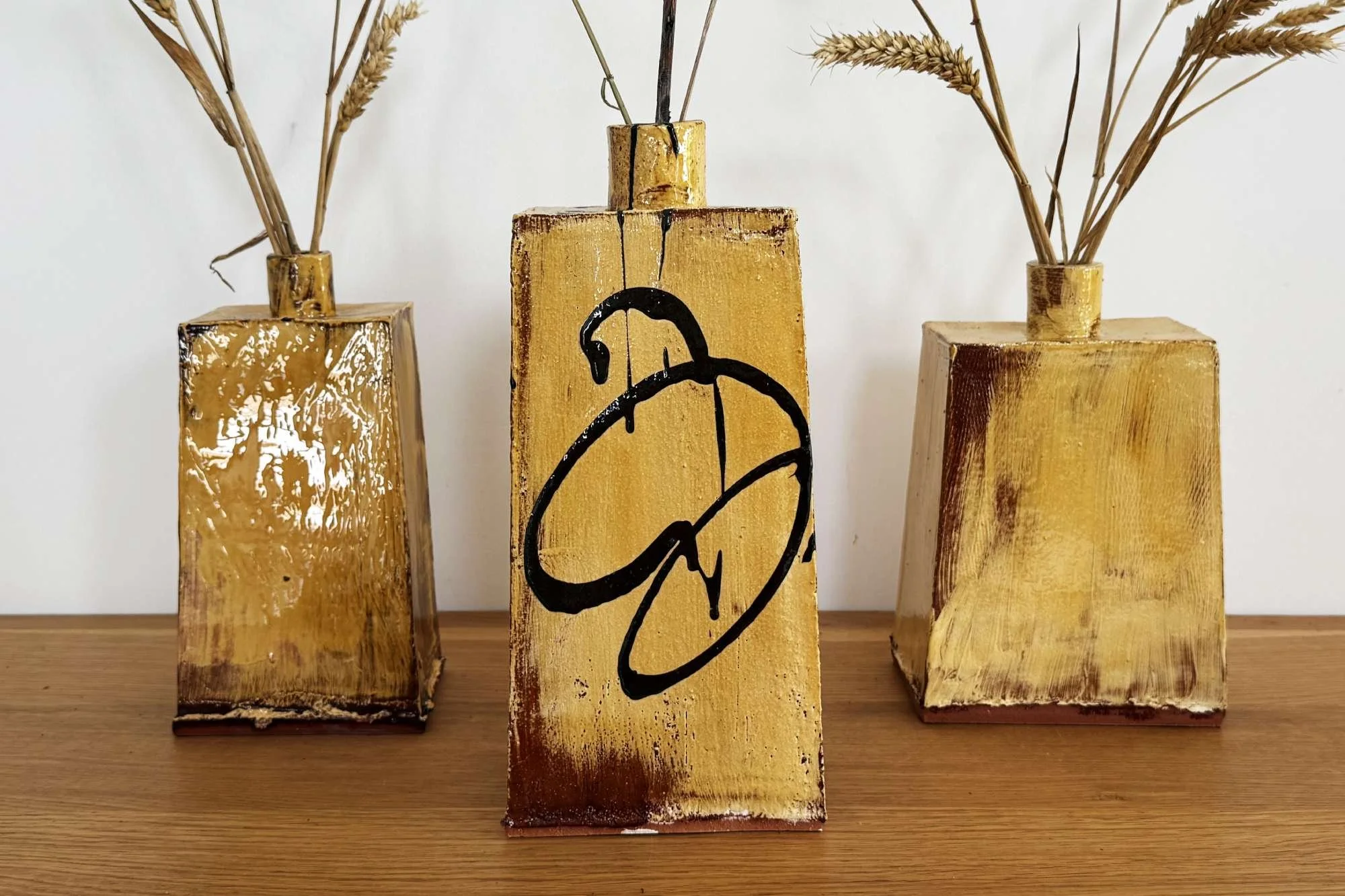 Three yellow ceramic vases with dried wheat and grass stems, placed on a wooden surface against a white background. The middle vase features a black abstract spiral design.