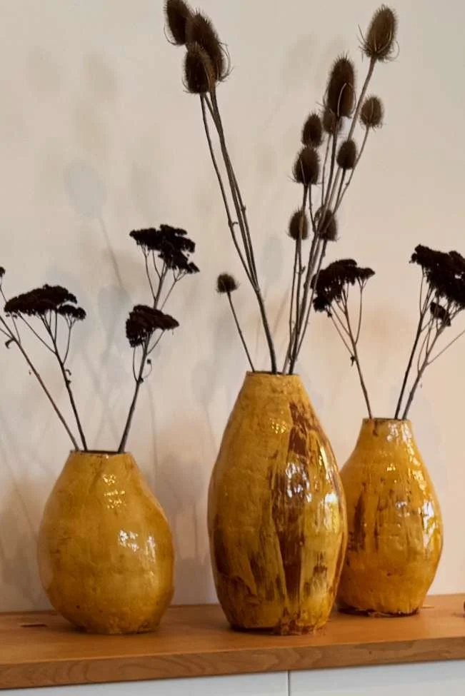 Three yellow ceramic vases with dried plants on a wooden surface