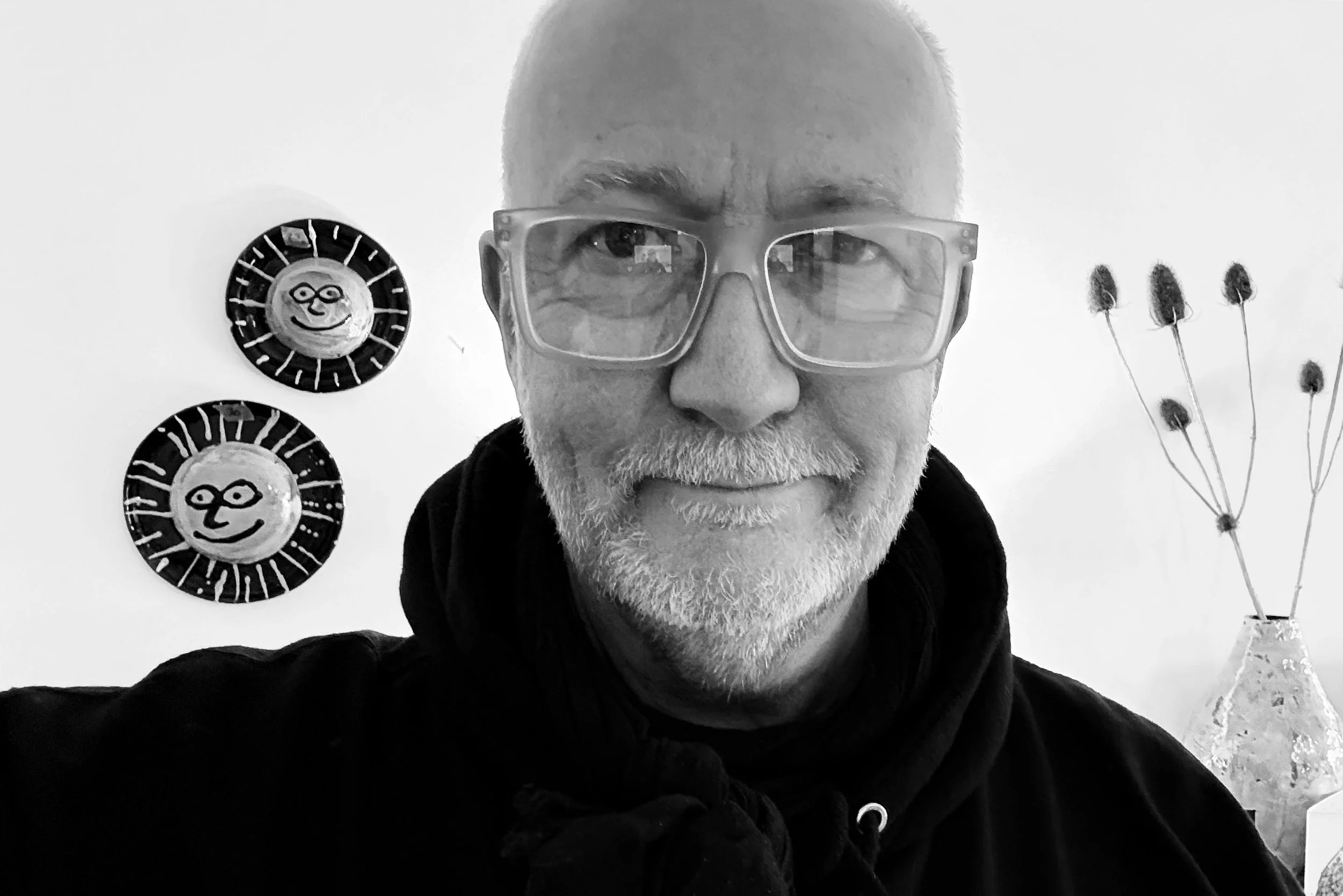 A Black and white photo of a smiling man with a beard and glasses, wearing a hoodie, standing indoors with wall decorations including two round wall hangings with abstract faces and a vase with tall, thin plants.