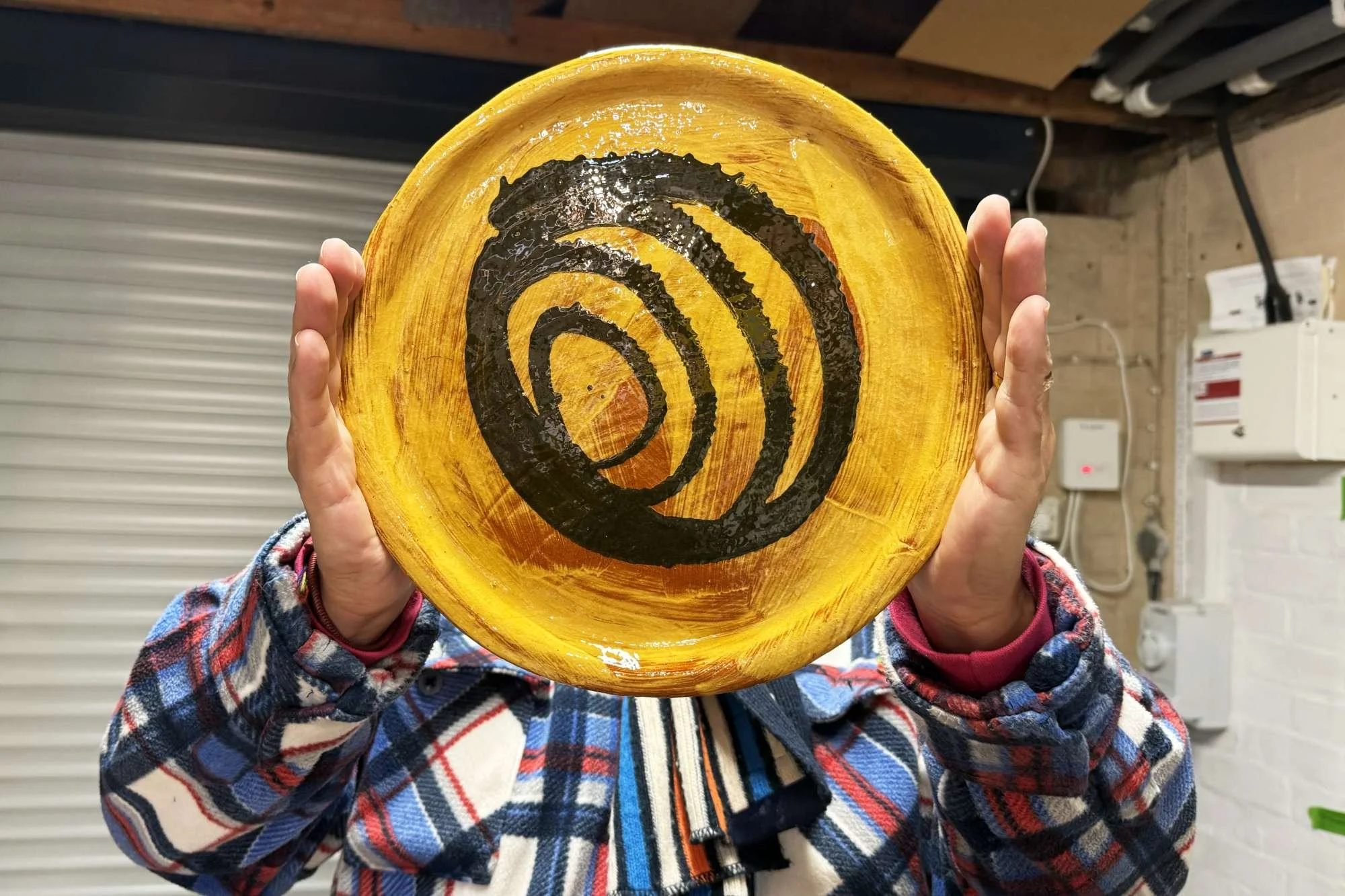 Alex Nisbett holding a round yellow glazed ceramic plate with a black spiral design at the centre, in his workshop.