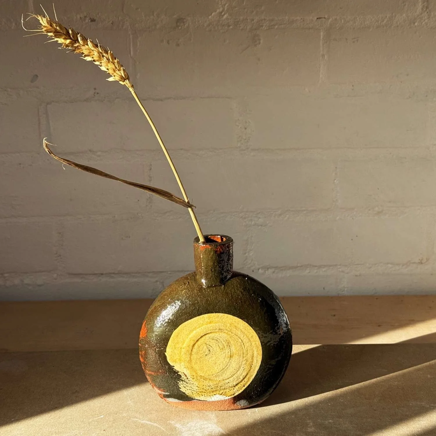 A small ceramic vase with a circular yellow wood grain pattern on the front, holding two dried wheat and grass stalks, placed on a wooden surface near a wall with shadow cast on the right side