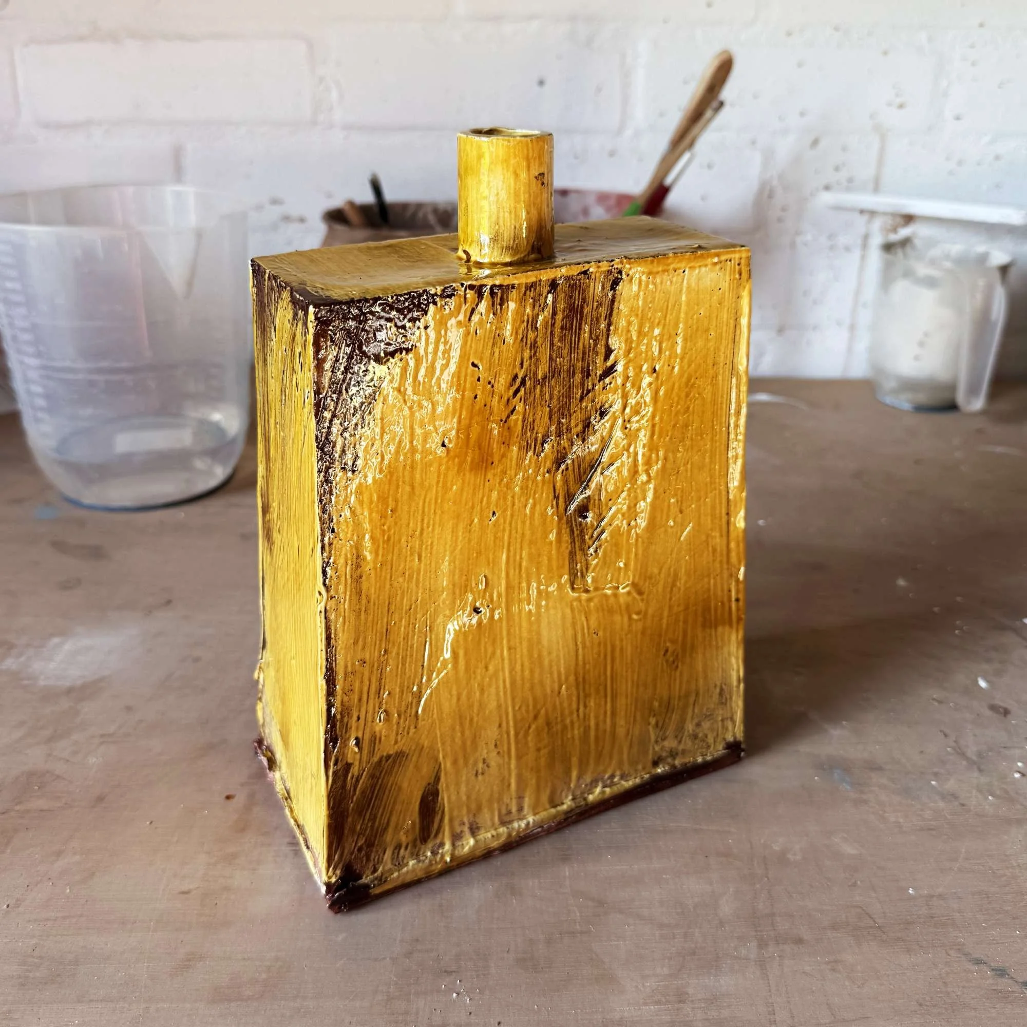 A rectangular wooden block with a small cylindrical piece on top, painted in yellow with some charred areas, positioned on a wooden work surface with tools and containers in the background.