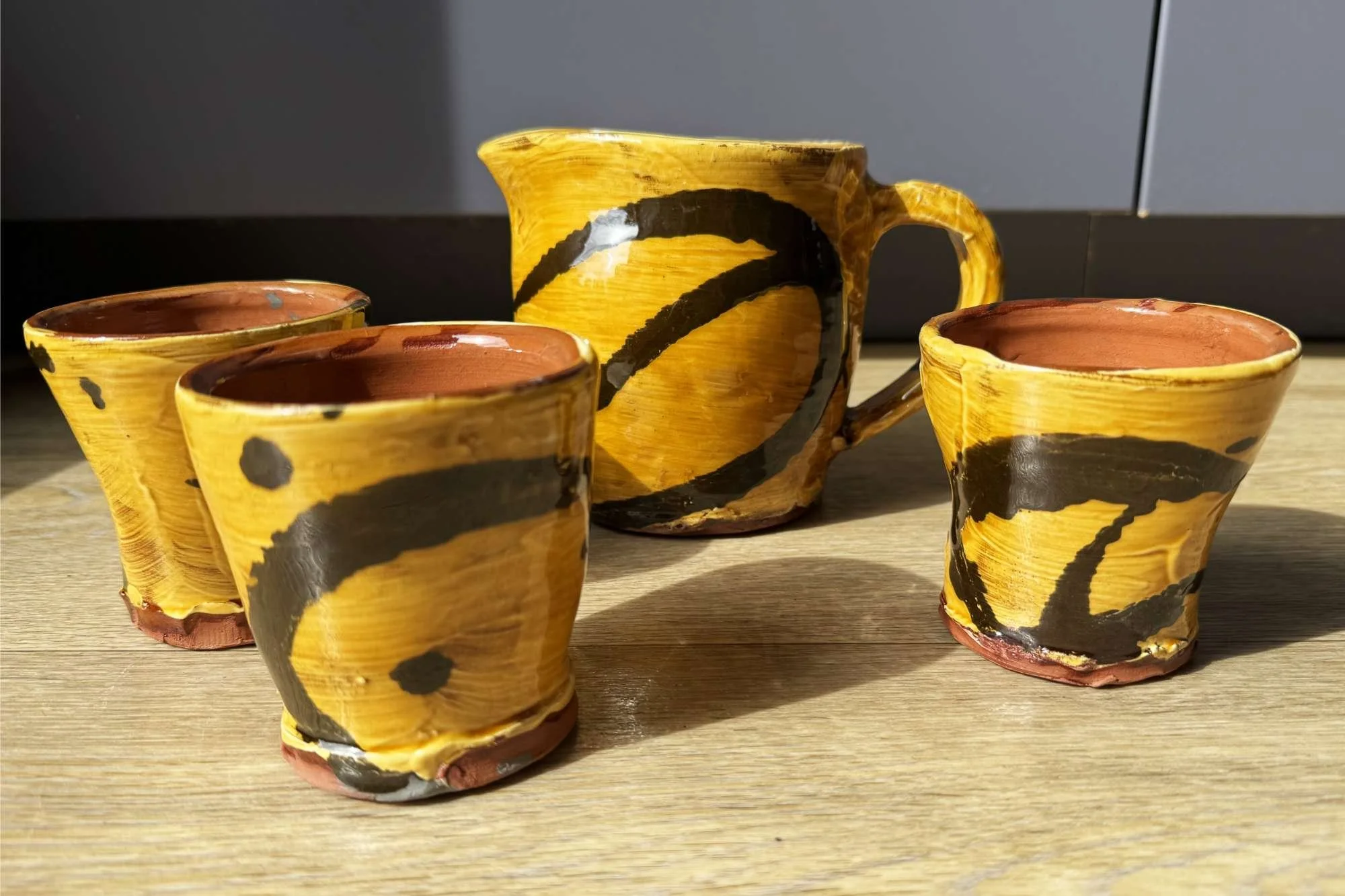 Set of four handmade ceramic cups and jug with a yellow background and black abstract lines, placed on a wooden surface.