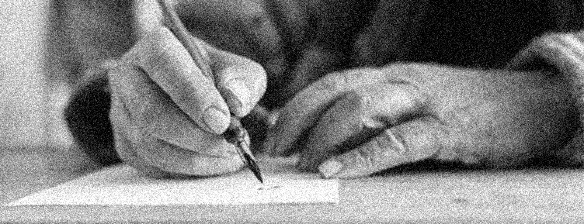 Close-up of a person writing with a pen on a piece of paper, capturing the hands and pen in black and white.