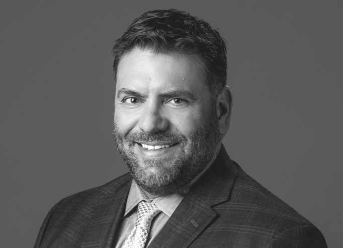 A smiling man with dark hair and a beard, wearing a suit and tie, in a black-and-white portrait.