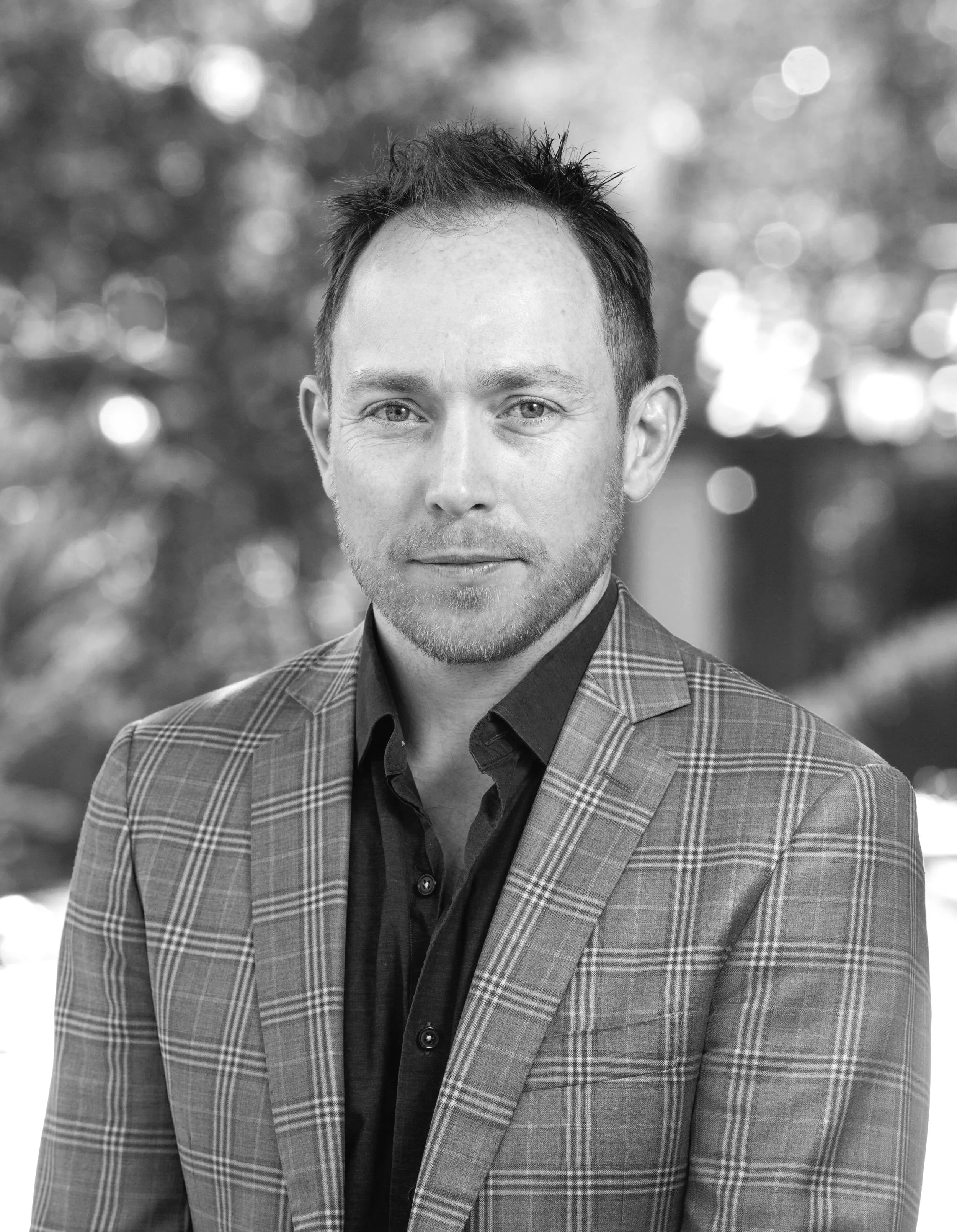 Black and white portrait of a man wearing a plaid blazer and a dark shirt, with a blurred background of trees and sunlight.