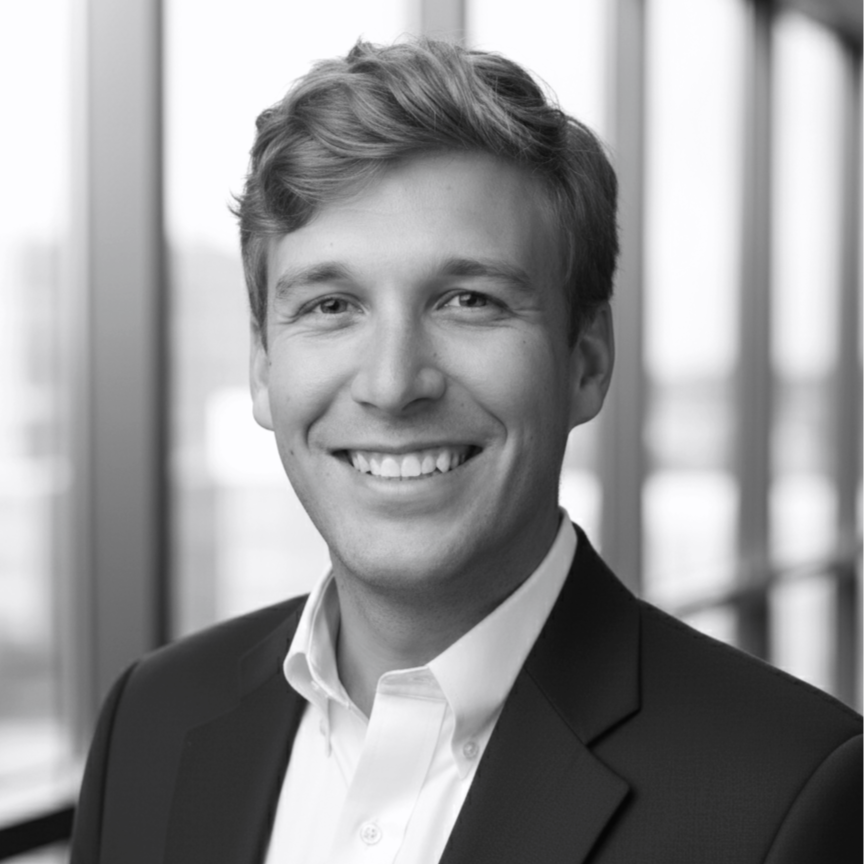 Black and white professional headshot of a young man with short wavy hair, smiling, wearing a suit jacket and white shirt, standing in front of large windows.