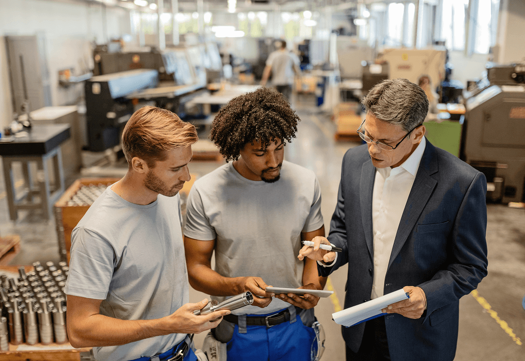 Three men in a manufacturing or factory setting having a discussion, looking at documents and a tablet, with machinery and tools in the background.