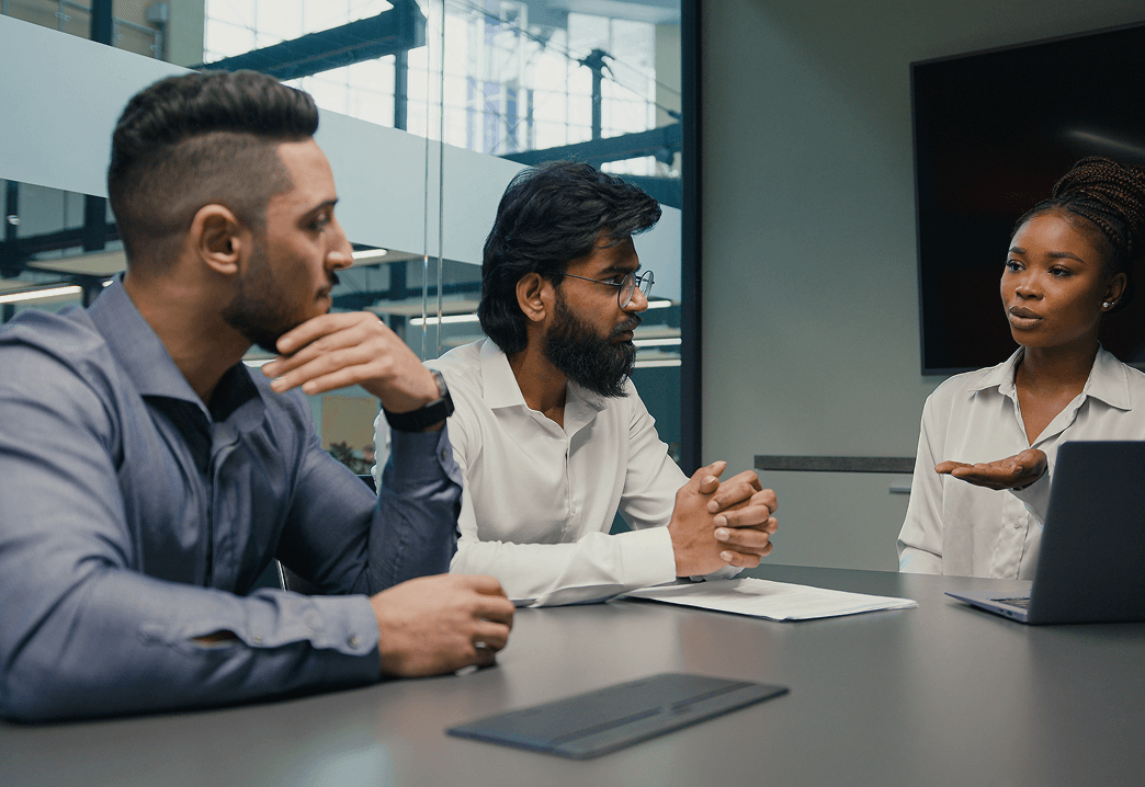 Three people in business attire participating in a discussion in a modern conference room with glass walls.