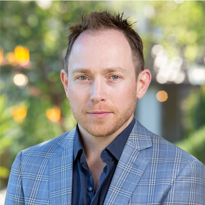 A man in a blue plaid suit jacket with a dark blue shirt standing outdoors with blurred trees and sunlight in the background.