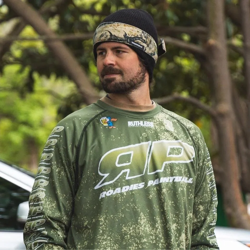 A man wearing a black beanie and camouflage neck gaiter, standing outdoors with trees in the background, wearing a green ARP Roadies Paintball jersey.