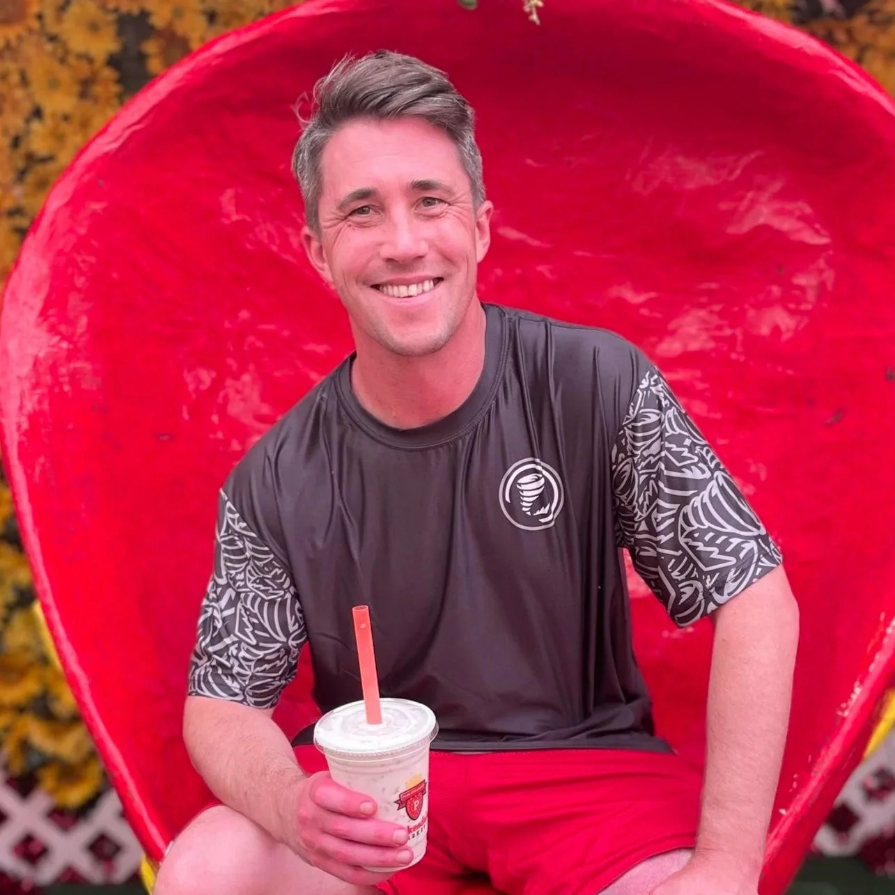 A man smiling while sitting in a red chair shaped like a heart background, holding a cup with a straw.