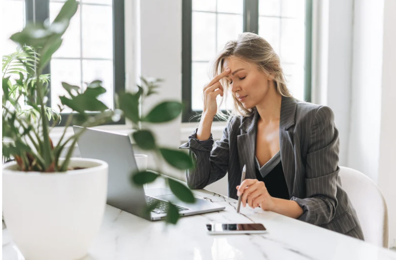 A woman sitting at a desk with a laptop, holding her forehead in frustration or stress, in an office with large windows and green plants.