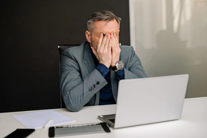 Frustrated professional at his desk in a gray suit putting his hands in his face.