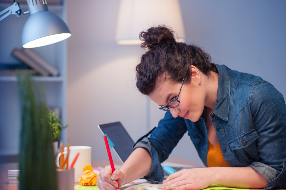 A woman with glasses, a jean jacket, and her hair in a bun diligently working on creating something at her desk