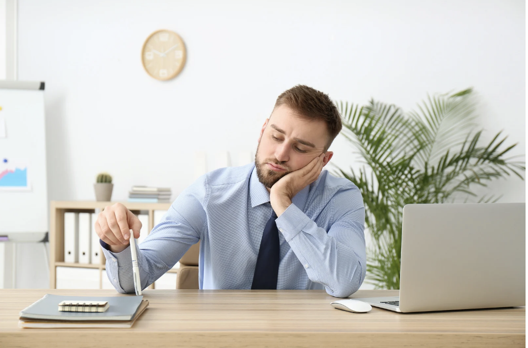 A tired businessman resting his head on his hand while sitting at a desk with a laptop, notebook, and pen in an office setting.