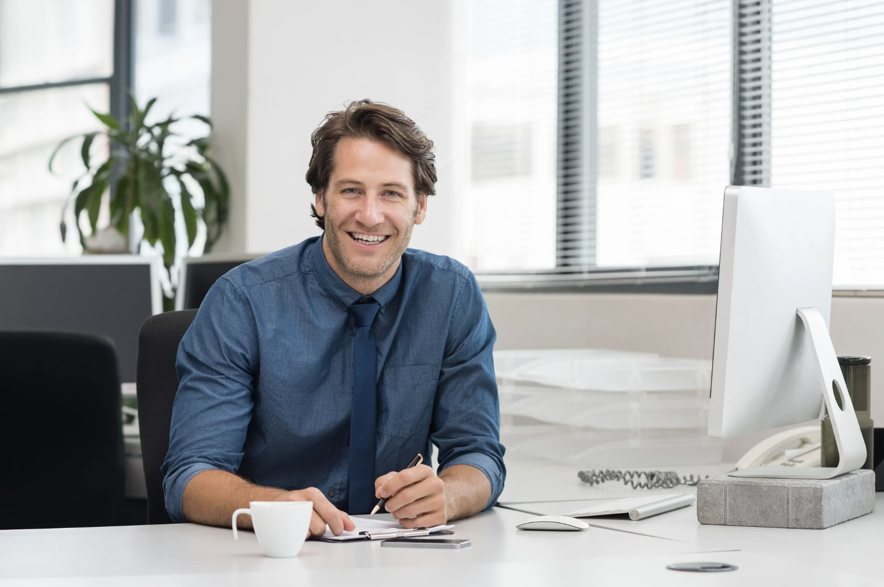 A smiling man in a blue shirt and tie sitting at a desk with a computer, coffee cup, and notepad in an office with large windows.