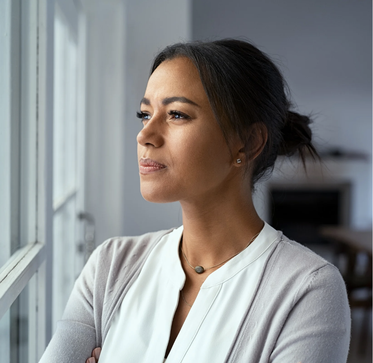 A woman with dark hair in a bun looking out a window, wearing a white top, gray cardigan, and necklace, in a well-lit indoor setting.
