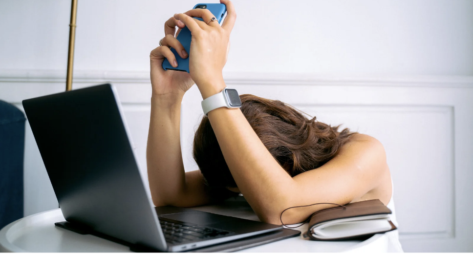 A woman with brown hair resting her head on her arms on a table, holding a phone above her head, with a laptop, notebook, and smartwatch present.