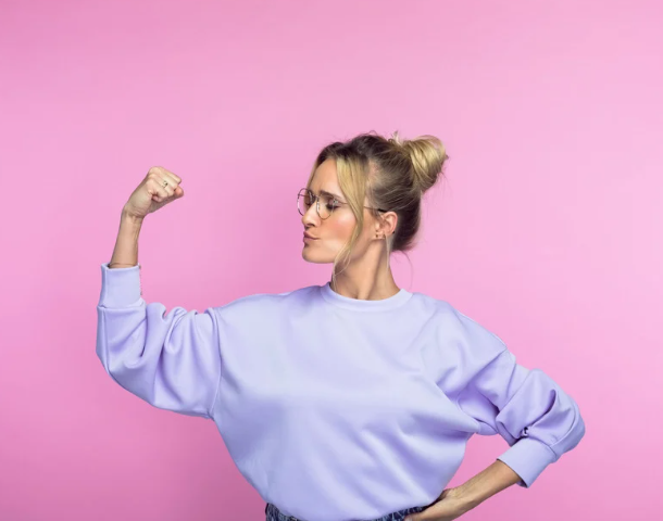 A blonde woman with glasses and a violet sweatshirt flexing her muscles
