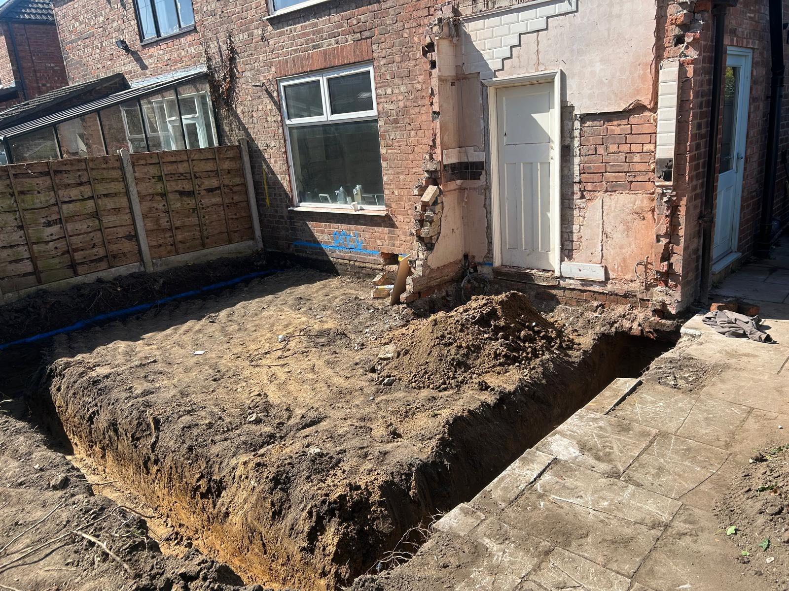 Construction site in backyard of brick house showing excavated ground, with dug-out area next to the house for renovation or foundation work, and tools and materials nearby.