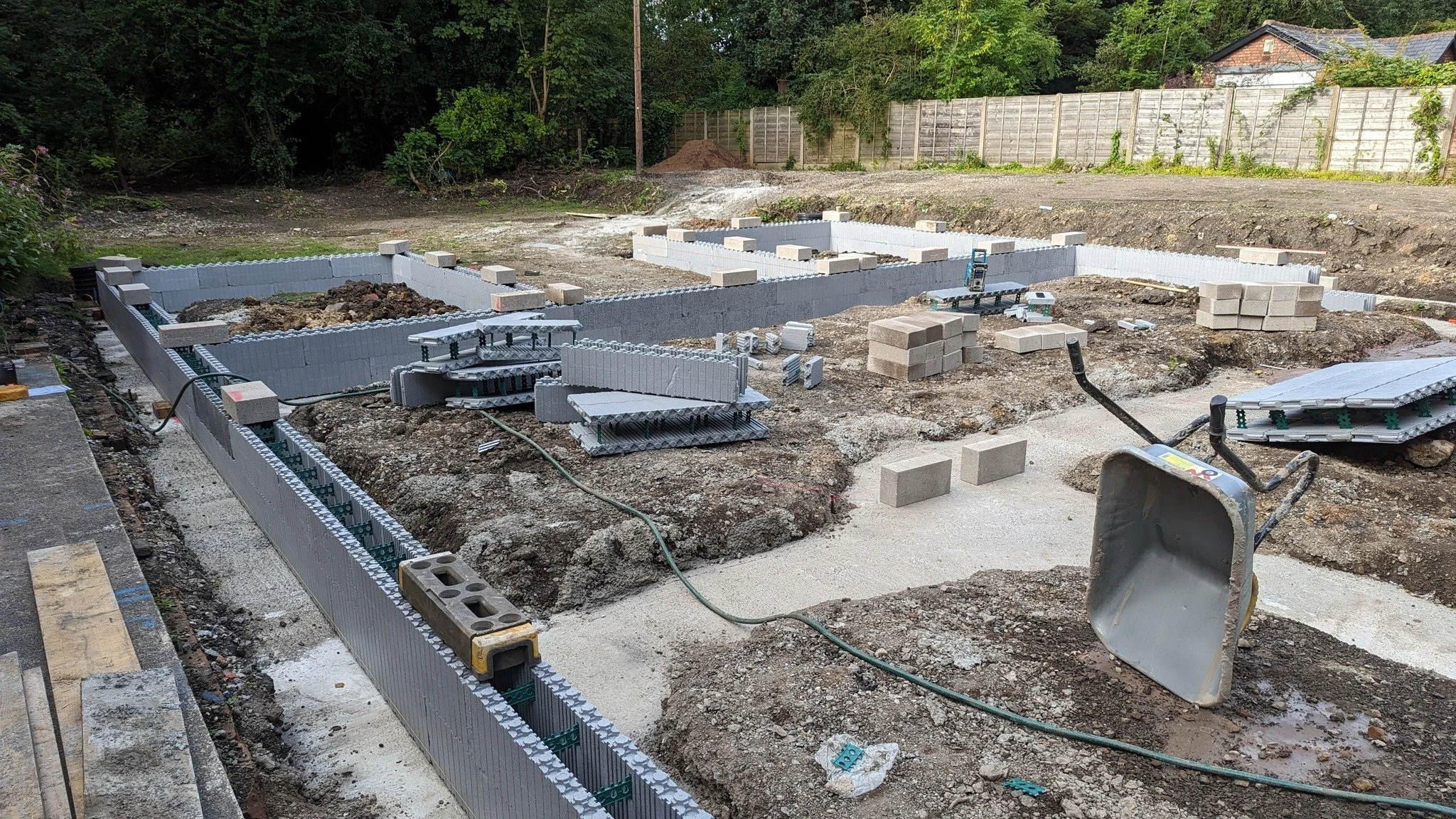 Construction site with building foundation in progress, concrete blocks, tools, and equipment on dirt ground surrounded by trees and fencing.