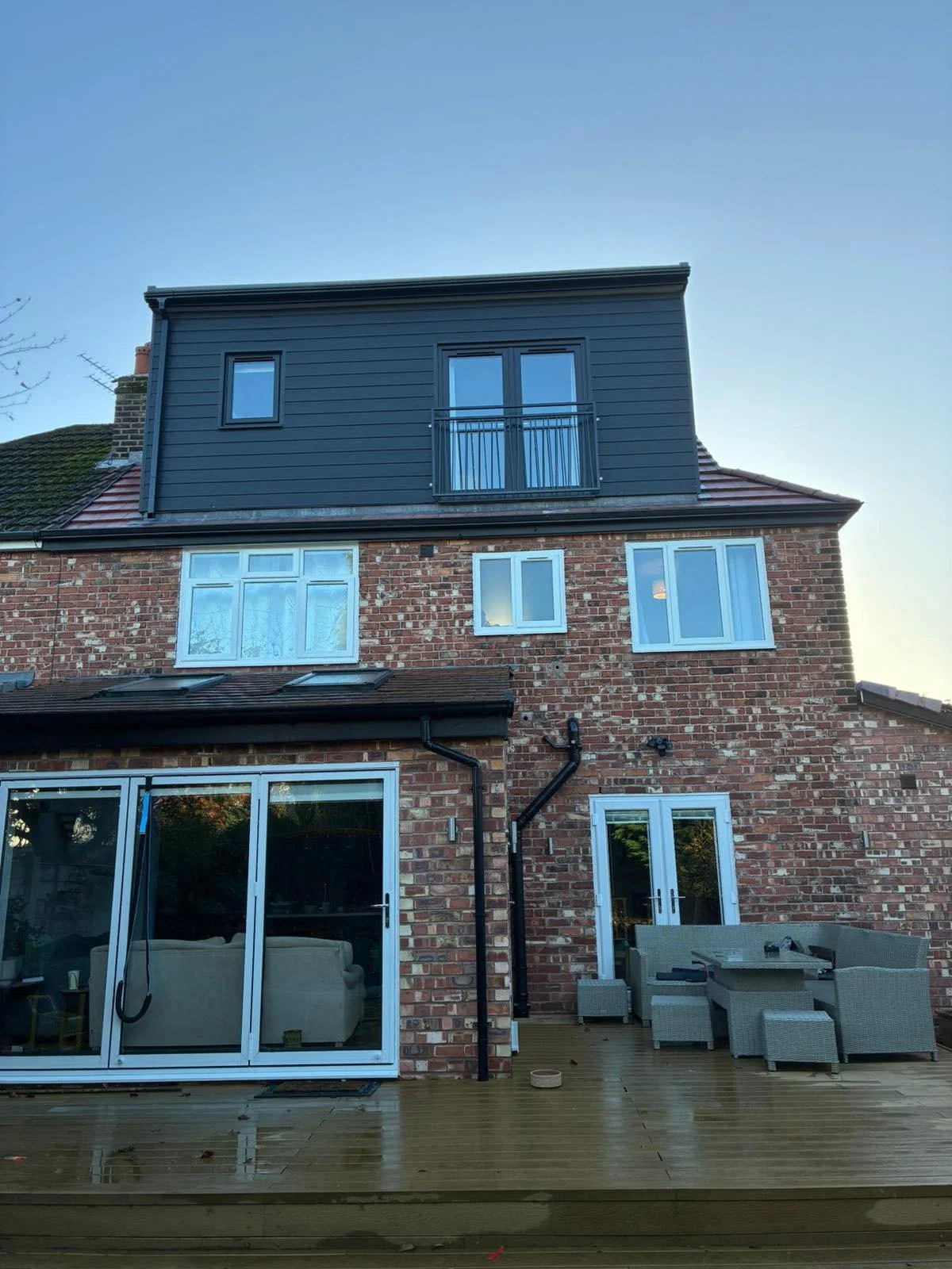 Backyard deck area of a brick house with sliding glass doors, outdoor furniture, and black gutters, with a second-story addition with dark siding and windows.