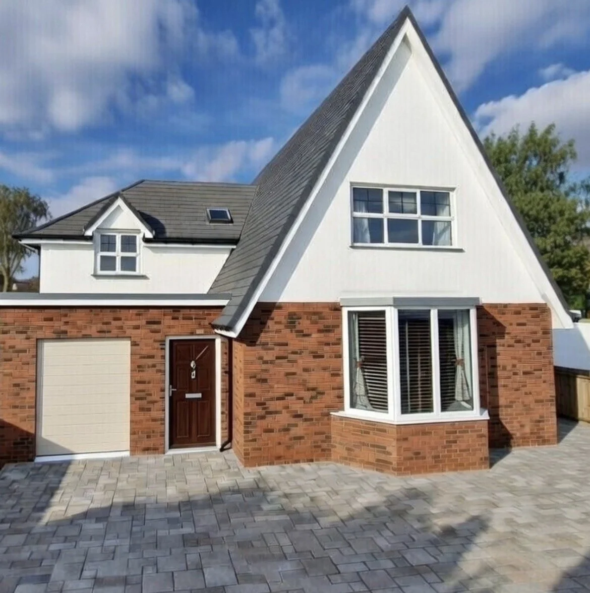 A house with a brick base and white upper part, featuring a unique steep triangular roof, a bay window with shutters, a small window on the dormer, and a garage door, with a paved driveway and a partly cloudy sky in the background.