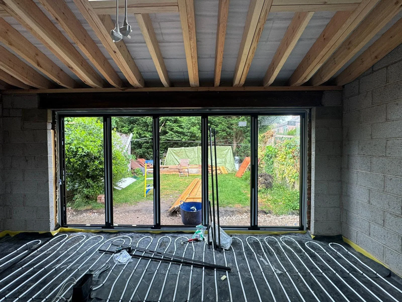 Interior of a room under construction showing a large window with black frames, a brick wall on the right, an unfinished ceiling with wooden beams, and underfloor heating pipes installed on a black vapor barrier.